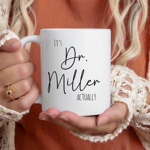 May include: A white ceramic mug with the text "It's Dr. Miller Actually" in black script. The mug is held by a person wearing a gold ring and a cream-colored sweater.