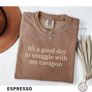 May include: Espresso-colored t-shirt with the text "it's a good day to snuggle with my cavapoo" in white. A silver necklace with a pendant rests on the shirt. The shirt is a Comfort Colors brand.