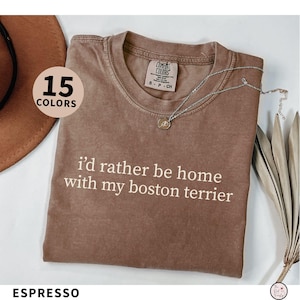 May include: Espresso-colored t-shirt with the text "I'd rather be home with my boston terrier" in white. The shirt has a crew neck and short sleeves. A silver necklace with a pendant is draped on the shirt. The image also shows a hat and dried plants.