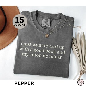 May include: A grey Comfort Colors t-shirt with the text "i just want to curl up with a good book and my coton de tulear." A gold necklace and a brown hat are also visible. The shirt is labelled "Pepper."