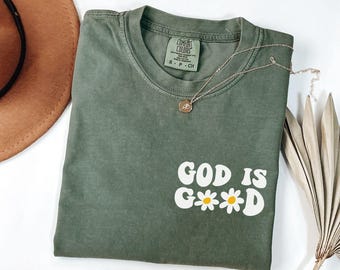 May include: A moss green t-shirt with the words "GOD IS GOOD" in white, with daisy accents. The shirt has a crew neck and short sleeves. A gold necklace with a pendant is draped on the shirt. A brown hat and dried plant are in the background.