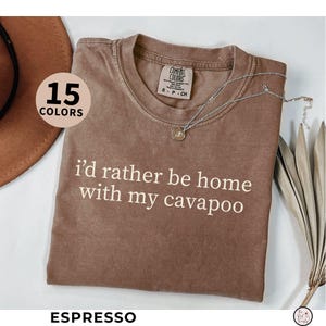 May include: Espresso-colored t-shirt with the text "I'd rather be home with my cavapoo" in white. The shirt has a crew neck and short sleeves. A silver necklace with a pendant is draped across the shirt. The image also shows a hat and dried plants.