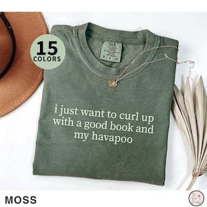 May include: Moss green t-shirt with the text "i just want to curl up with a good book and my havapoo" in white. The shirt has a crew neck and short sleeves. A gold necklace and a brown hat are also visible.