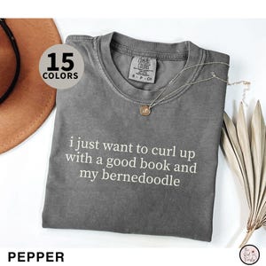 May include: A gray t-shirt with the text "i just want to curl up with a good book and my bernedoodle" in white. The shirt is displayed with a necklace and a hat. The word "PEPPER" is at the bottom.