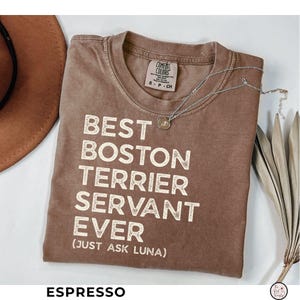 May include: Espresso-colored t-shirt with the text "BEST BOSTON TERRIER SERVANT EVER (JUST ASK LUNA)" in white block letters. A silver necklace with a small pendant rests on the shirt. The shirt is a Comfort Colors brand.