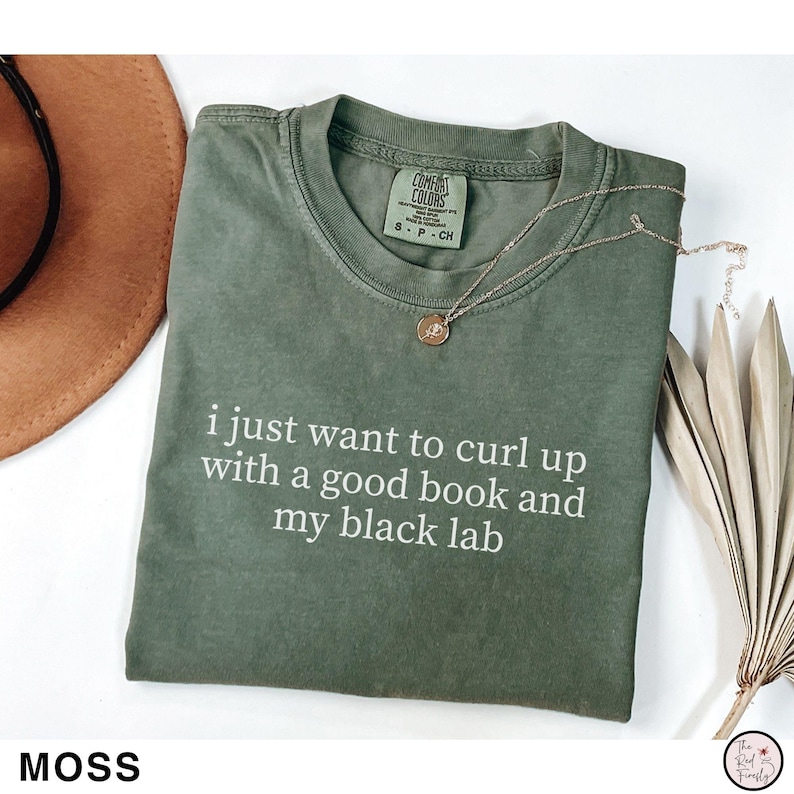 K&ouml;nnte beinhalten: Ein moosgr&uuml;nes T-Shirt mit dem wei&szlig;en Text "i just want to curl up with a good book and my black lab". Eine goldene Halskette und ein brauner Hut sind ebenfalls sichtbar.