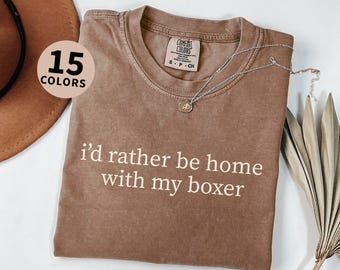 May include: Espresso-colored t-shirt with the text "I'd rather be home with my boxer" in white. The shirt has a crew neck and a Comfort Colors tag. A silver necklace with a pendant is draped across the shirt. The image also shows a hat and dried plants.