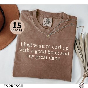 May include: Espresso-colored t-shirt with the text "i just want to curl up with a good book and my great dane." The shirt has a crew neck and short sleeves. A silver necklace with a pendant is draped across the shirt.