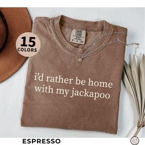 May include: Espresso-colored t-shirt with the text "I'd rather be home with my jackapoo" in white. The shirt has a crew neck and short sleeves. A silver necklace with a pendant is draped across the shirt. The words "15 COLORS" are in a circle.