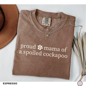 May include: An espresso-colored short-sleeve t-shirt with a crew neckline. The shirt features the white text "proud mama of a spoiled cockapoo" with a paw print icon. A silver-toned necklace with a small circular pendant rests on the shirt.