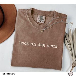 May include: An espresso-colored short-sleeve t-shirt with the text "bookish dog mom" printed in white across the chest. The shirt features a crew neckline and a Comfort Colors brand label on the inside collar, indicating a size small.