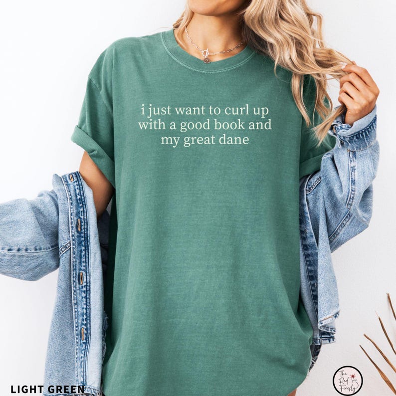 May include: Light green t-shirt with the text "i just want to curl up with a good book and my great dane" in white. The shirt has rolled-up sleeves and a crew neck. A denim jacket is draped over the arm.