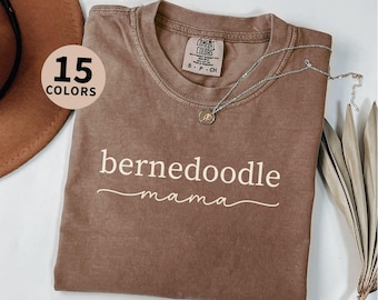 May include: Espresso-colored t-shirt with the words "bernedoodle mama" in white script. The shirt has a crew neck and short sleeves. A silver necklace with a pendant rests on the shirt. The image also shows "15 COLORS" in a circle.