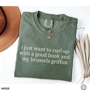 May include: A moss green short-sleeve t-shirt with a crew neckline and a Comfort Colors label. The shirt features white text that reads: i just want to curl up with a good book and my brussels griffon. A thin gold-tone chain necklace is placed on top.
