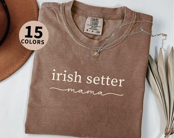 May include: Espresso-colored t-shirt with the text "irish setter mama" in white script. The shirt has a crew neck and short sleeves. A silver necklace with a pendant rests on the shirt. The image also shows a hat and dried plants.