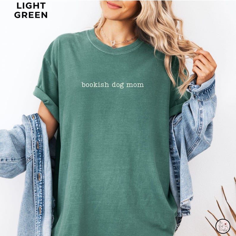 May include: A light green short-sleeved t-shirt featuring the white text "bookish dog mom" printed across the chest. The garment has a crew neckline and a relaxed fit, layered under a light blue denim jacket with visible metal buttons.