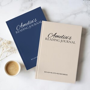 May include: Two hardcover reading journals, one navy blue and one beige, both featuring the text Amelia's READING JOURNAL in elegant script and serif fonts, with the phrase READ+RATE+REMEMBER printed in black at the bottom of the beige cover.