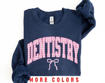 May include: Navy blue sweatshirt with the word "DENTISTRY" in pink gingham lettering. A pink bow is below the word. The text "MORE COLORS" is in red, and "NAVY" is in white. The sweatshirt is folded.