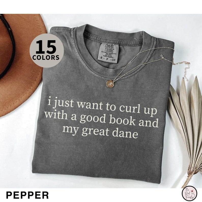 May include: Pepper-colored t-shirt with the text "i just want to curl up with a good book and my great dane." The shirt has a crew neck and short sleeves. A gold necklace is draped across the shirt.