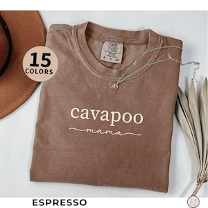 May include: Espresso-colored t-shirt with the words "cavapoo mama" in white script. The shirt has a crew neck and short sleeves. A silver necklace with a pendant rests on the shirt. The words "15 COLORS" are in a circle on the left.