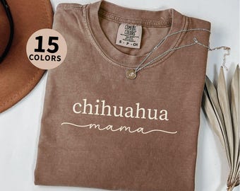 May include: Espresso-colored t-shirt with the words "chihuahua mama" in white script. The shirt has a crew neck and a Comfort Colors tag. A silver necklace with a pendant is draped on the shirt. The image also shows a hat and dried plants.