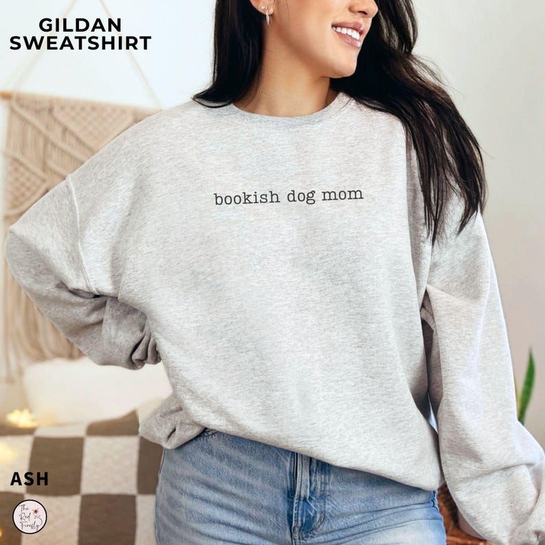 May include: A light gray crewneck sweatshirt featuring the text "bookish dog mom" printed in black on the chest. The garment has long sleeves, a ribbed neckline, and a relaxed fit, paired with blue denim jeans. The text "GILDAN SWEATSHIRT" and "ASH" are visible in the corners.