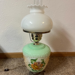 May include: A vintage table lamp with a white glass shade and a green base. The base features a hand-painted floral design with yellow and orange roses. The lamp has a brass-colored metal accent and a clear glass chimney.