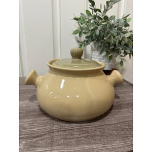 May include: A tan ceramic casserole dish with a lid. The dish has two side handles and a rounded shape. The lid is a lighter shade of tan and has a small knob. The dish is sitting on a wooden surface.