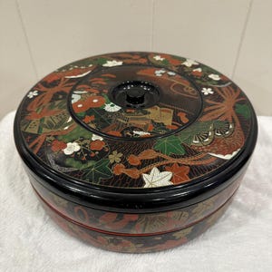May include: A round, black lacquered box with a lid, featuring intricate floral and landscape designs in red, green, and gold. The box has a black knob on top and a red accent band around the base. The box is sitting on a white surface.