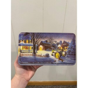May include: Rectangular metal tin with a winter farm scene. The image depicts a house, barn, tractor, and dog in a snowy landscape under a starry sky. The tin is held by a hand.