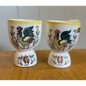 May include: Two white ceramic egg cups with a rooster design. The cups have a yellow rim and a decorative rooster illustration in red, green, and yellow. The base features a floral design. The cups are on a wooden surface.