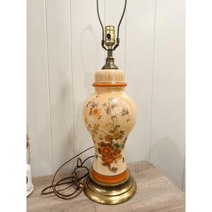May include: A vintage table lamp with a beige ceramic base and a brass-colored metal base. The lamp features a floral design in orange, yellow, and brown. The lamp is approximately 20 inches tall.