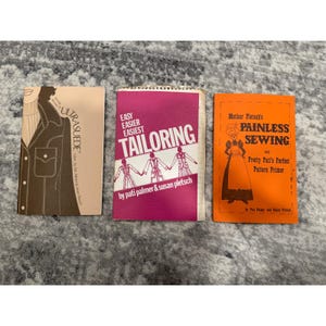 Pode incluir: Três livretos de padrões de costura vintage. Os livretos são intitulados "Ultrasuede", "Easy Easier Easiest Tailoring" e "Mother Pletsch's Painless Sewing". O livreto "Tailoring" é magenta e branco. O livreto "Painless Sewing" é laranja.