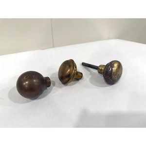 May include: Three round metal door knobs with a weathered finish. One knob is dark brown, one is a smooth brass tone, and the third is a textured, tarnished brass color featuring a square metal spindle extending from the back of the knob.