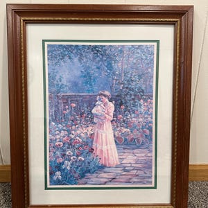 May include: Framed print of a woman in a pink dress holding a baby in a garden. The artwork features a brick pathway, lush flowers in shades of pink, purple, and white, and a dark brown wooden frame with a gold inner border.