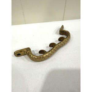 May include: A curved brass drawer pull featuring a textured, scale-like pattern along its surface. The handle has two flat mounting ends with pre-drilled holes and three small, rounded decorative protrusions extending from the inner curve.