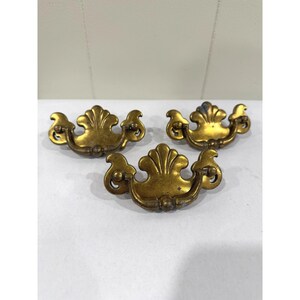 Peut inclure: Trois poignées de tiroir en métal doré, dotées d'une plaque arrière décorative en forme de coquille et d'une poignée incurvée mobile. Chaque pièce présente une finition vieillie et patinée avec des variations de couleur et de texture.