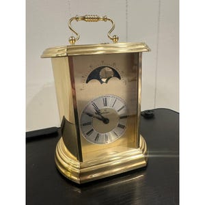 May include: A gold-toned mantel clock with a handle and a round base. The clock face has Roman numerals and a moon phase indicator. The clock is rectangular with a curved top and sides.