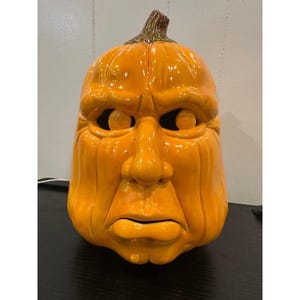 Puede incluir: Decoración de Halloween con forma de calabaza de cerámica con una cara de mal humor. La calabaza es de color naranja vibrante con una superficie texturizada y un tallo marrón. Los ojos, la nariz y la boca están tallados en la calabaza, dándole un aspecto único y expresivo.