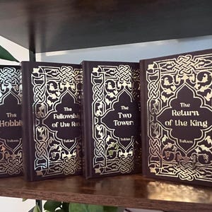 May include: A collection of four hardcover books with dark brown covers and gold foil lettering. The titles include "The Hobbit," "The Fellowship of the Ring," "The Two Towers," and "The Return of the King."