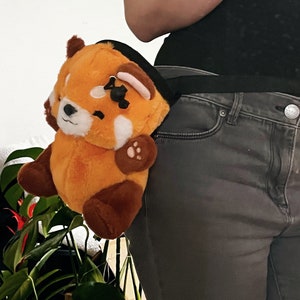 Puede incluir: Un panda rojo de peluche con un parche negro en el ojo está sujeto a un cinturón negro. El panda rojo lleva una camisa a rayas blancas y negras. El cinturón se lleva alrededor de la cintura de una persona que lleva pantalones vaqueros grises oscuros.