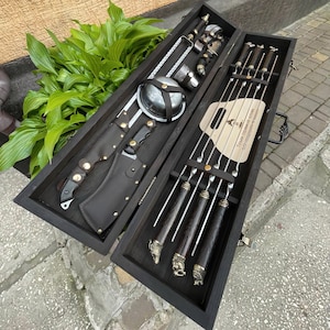 May include: A wooden box with a hinged lid, containing a set of metal skewers and a large knife with a leather sheath. The skewers have wooden handles and are arranged in a row. The box is open and the skewers are visible. The text on the box reads "Товары с доставкой по России".