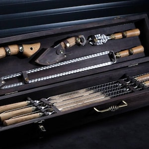 Puede incluir: Un juego de caja de madera oscura que contiene herramientas para asar a la parrilla. El juego incluye un hacha pequeña con funda de cuero, un cuchillo, un tenedor para carne, una hoja larga dentada y varias brochetas. Las herramientas tienen mangos de madera y están dispuestas dentro de la caja.