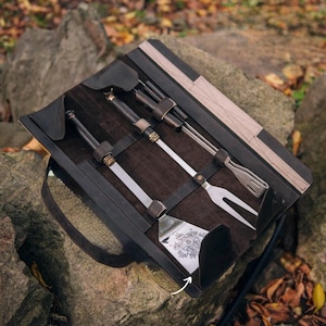 Puede incluir: Un estuche enrollable de cuero negro con un interior de gamuza marrón contiene un juego de cinco herramientas para asar a la parrilla. Las herramientas incluyen una espátula, pinzas, un tenedor, un pincel para untar y una herramienta de limpieza. El estuche tiene una correa para llevar.
