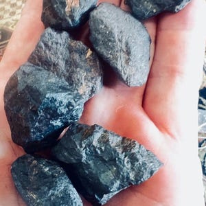 May include: A collection of six raw, dark charcoal-coloured stones with irregular, jagged shapes and textured surfaces. The stones exhibit a matte finish with subtle hints of metallic grey and brown mineral inclusions scattered across their uneven edges.