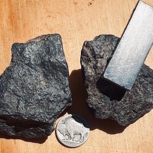 May include: Two irregular, dark, textured rocks are positioned next to a silver-coloured rectangular magnet and a United States five-cent nickel coin, which serves as a scale reference for the size of the mineral specimens.