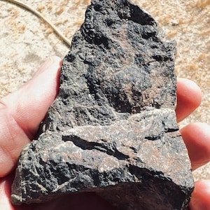Puede incluir: Una roca dentada de color gris oscuro y negro con una textura superficial rugosa e irregular. La piedra presenta bordes afilados y angulares con depósitos minerales visibles, mostrando una mezcla de tonos carbón y reflejos marrones.