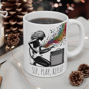 May include: White ceramic mug with a black and white illustration of a person playing an electric guitar with colorful musical notes. The text "Sip, Play, Repeat" is printed on the mug.