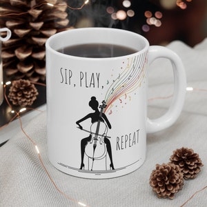 May include: White ceramic mug featuring a black silhouette of a person playing a cello. The mug has the text "Sip, Play, Repeat" and colourful musical notes.