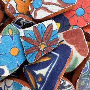 以下が含まれることがあります： 花柄のカラフルな手描きセラミックタイルのコレクション。タイルはさまざまな形とサイズで、青、赤、オレンジ、黄、緑の色が混ざり合っています。